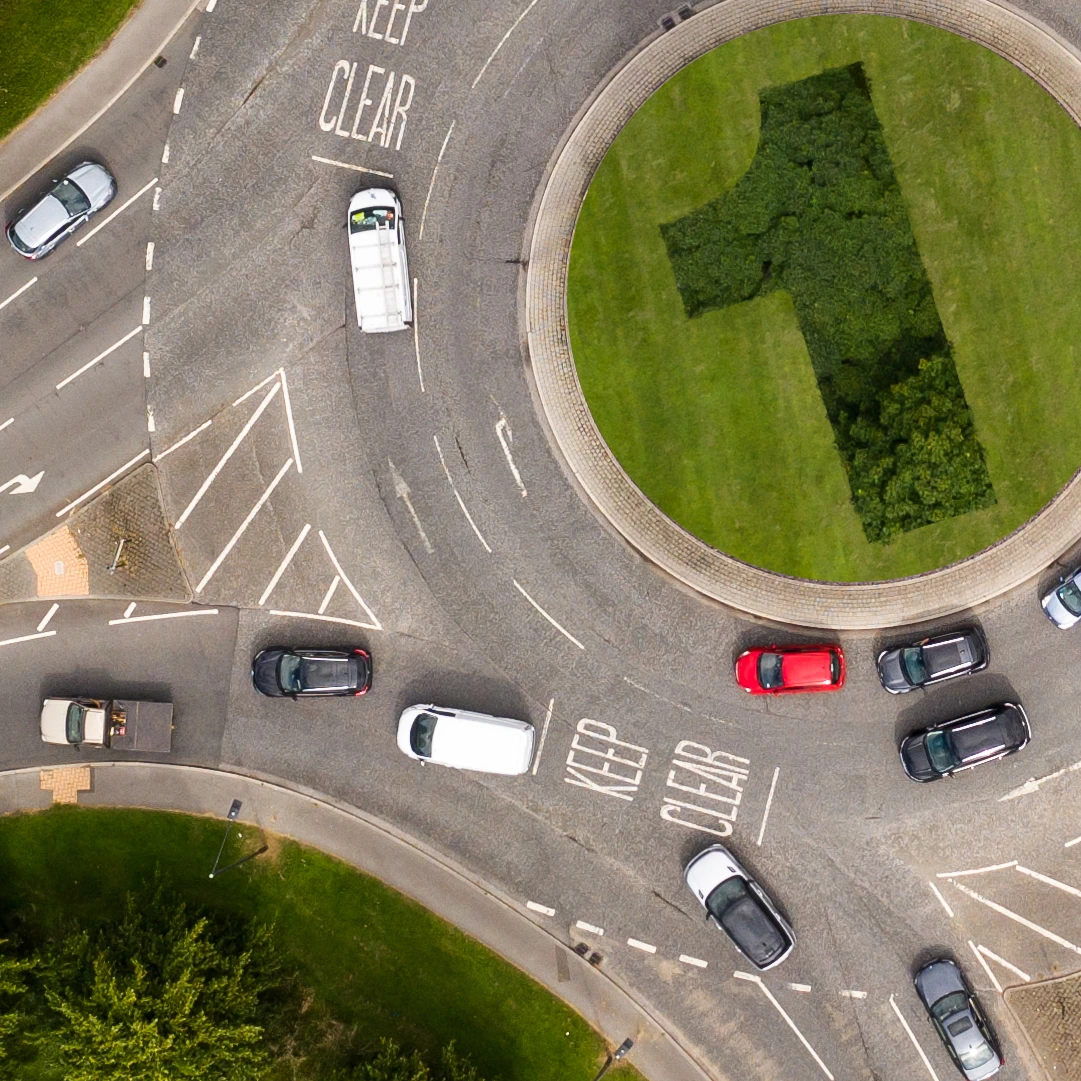A road roundabout from above. There are car going round the roundabout. In the centre of the roundabout is a green space, trees and grass, with a shape of a 1 showing